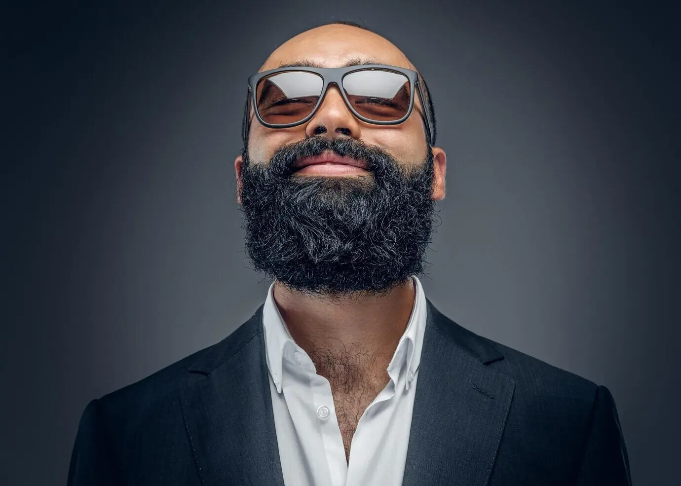 Porträt eines modischen bärtigen Mannes im Anzug und mit Sonnenbrille, freigestellt vor grauem Hintergrund.